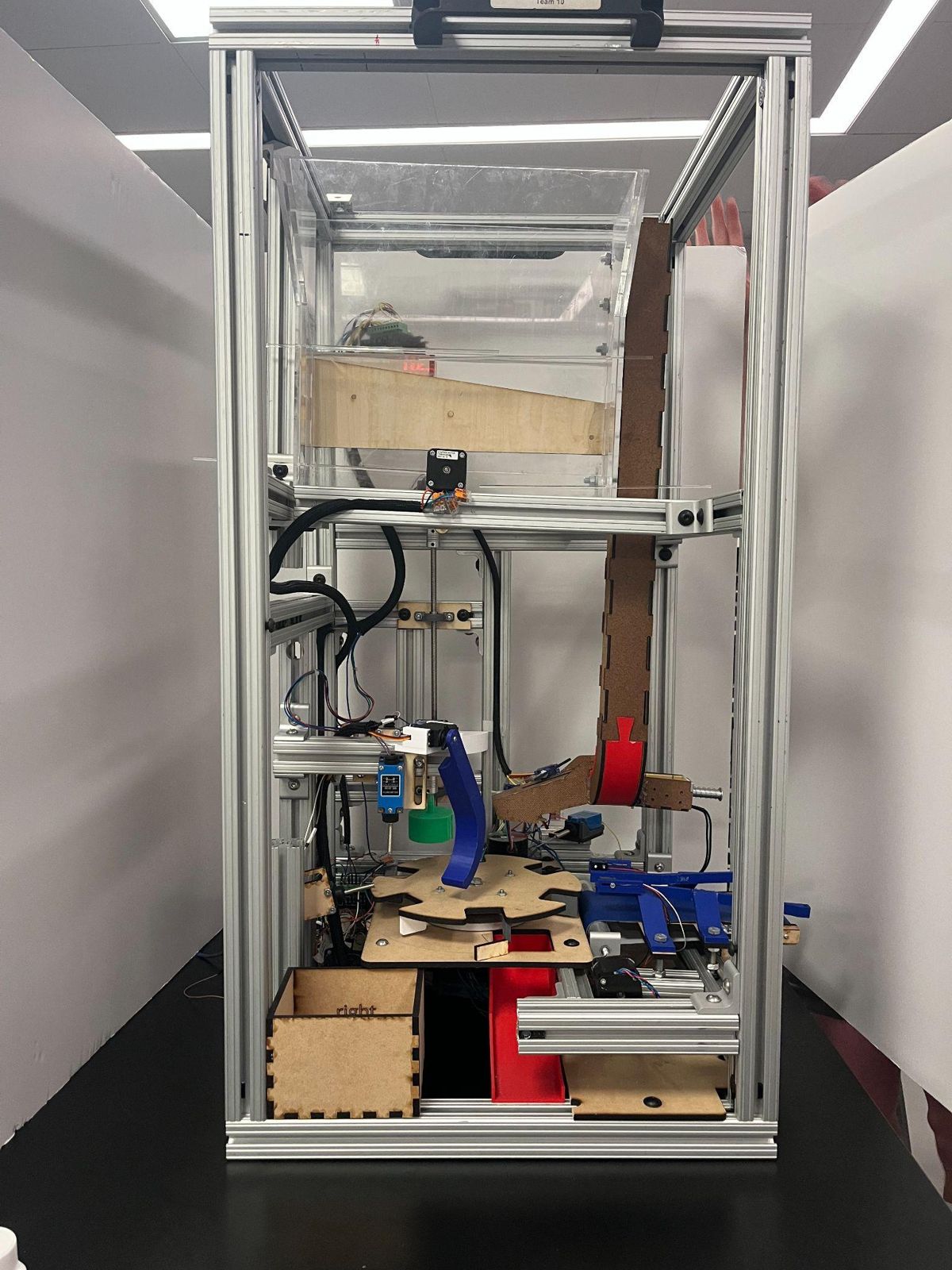 The complete capping machine inside a 26 by 20 by 40 inch aluminum 80/20 frame: acrylic cap descrambler on top, capping head and blue rejector arm above a laser-cut rotary table, conveyor on the right, collection bins below