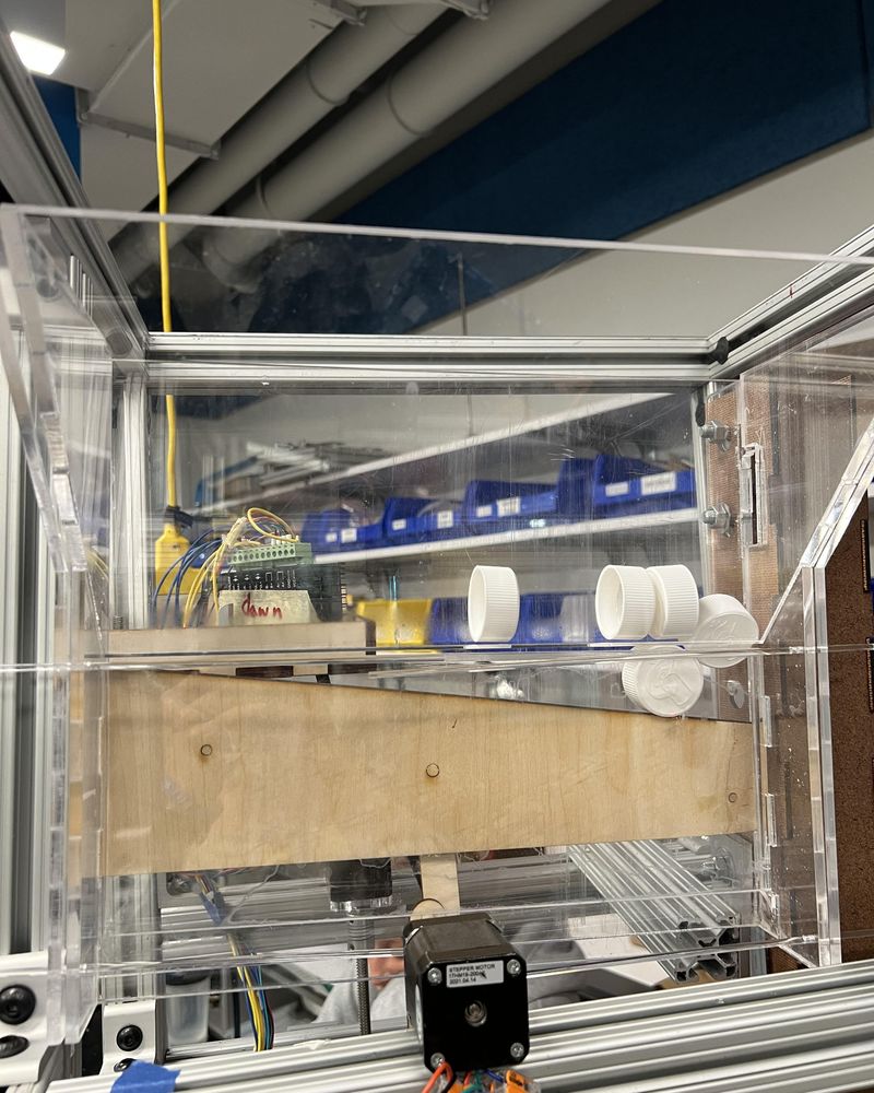 Clear acrylic descrambler hopper with loose white caps on an angled platform, stepper motor below
